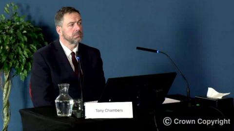 Tony Chambers, who has short grey hair and a short grey beard, sits at a black desk before a set of microphones, a computer screen and a jug of water. 