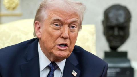 President Trump wears a blue suit, white shirt and blue tie with an American flag pin while speaking to reporters in the Oval Office, a bust of Winston Churchill is visible behind him..