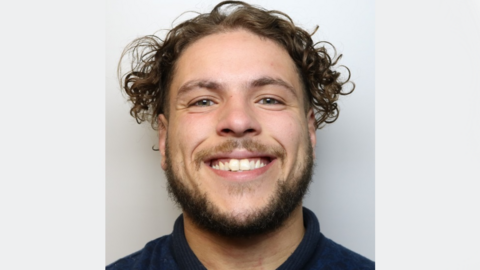 A police mugshot of Reuben Brown, wearing a dark blue polo shirt and standing against a grey background. He has curly brown hair and a beard, and is grinning at the camera.
