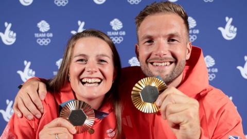 Emily Ford on the left - who has long brown hair - holding her bronze medal up to the camera; Tom Ford, who has a short, dark beard/stubble, with brown hair, is holding up his medal for the camera 