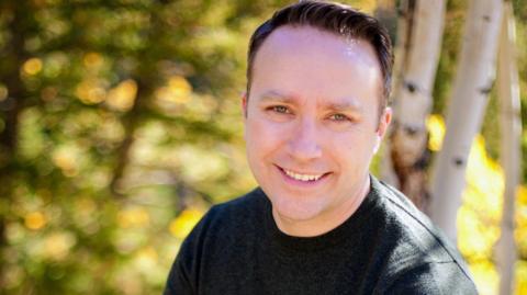 Greg Daily, wearing a black t-shirt, smiles at the camera, in front of trees