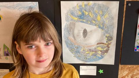 A young girl with long hair standing in front of a drawing of a dragon's eye.