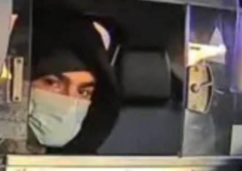 A hooded man wearing a face mask sits in the back of a vehicle looking through a dividing window