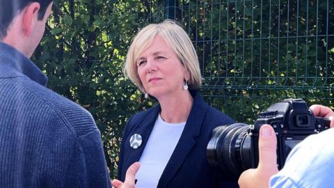 A woman with cropped blonde hair, wearing a blue blazer and white top, looking at a man wearing a blue jumper. An SLR camera is also pointing at her. Behind her is a metal fence with bushes growing behind it.