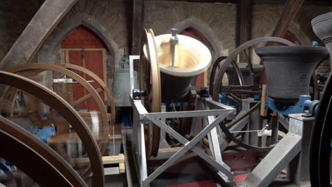 A bell being held up by metal. It is mid being rung so is moving towards the camera. There are other bells that are angles towards the sky to the right of it and being the main bell to the left is a bell facing downwards. Behind it is a wall with three doors. 