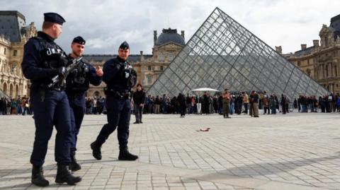 Three French police officers, one carrying a firearm, are seen walking on the square in front of the glass pyramid structure of the Louvre in Paris