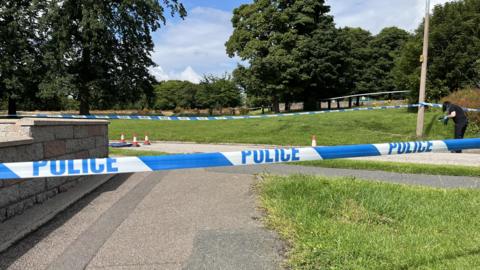 A strip of blue and white police tape across the image. There is a grey wall on the left hand side and a grey path going through the middle. A patch of green grass is on the right hand side. On the far right of the image, a police worker with long, dark hair and wearing dark clothing and blue protective gloves is partially bent over and is holding something in their hands.