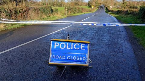 A two lane country road with hedgerows and green fields either side. A road closed police sign is on the road and a barrier is across the road. A back van can be seen at the bottom of the road.