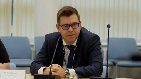 Nik Johnson is wearing a suit and tie and is sat behind a desk, with his hands clenched. A microphone is in front of him