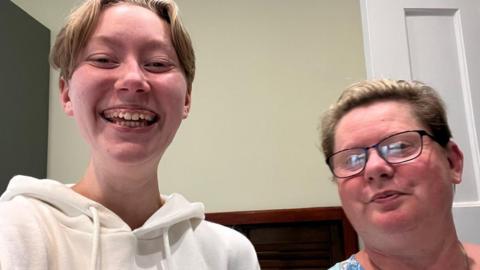 Chloe and Wendy smiling at the camera at home. Chloe has short blonde hair and Wendy has her hair tied back and is wearing dark rimmed glasses.