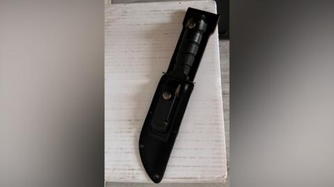A combat knife in a black holster is sat on what appears to be the top of an off-white table