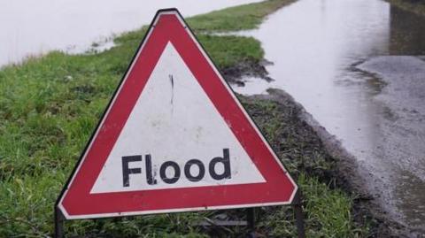 Stock image showing a flood warning sign on a grass verge next to a flooded lane.