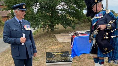 A man in a light blue RAF uniform and cap which is decorated with medals next to a piper carrying bag pipes. They are either side of the new memorial which has a black front with a picture of an aircraft and writing in gold. 