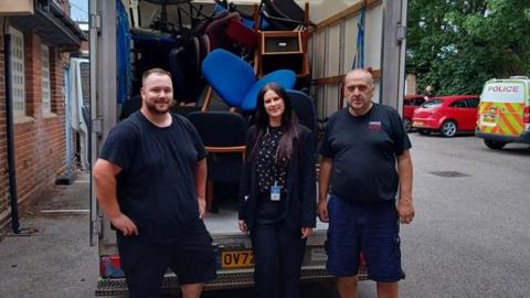 Two men stand either side of a woman at the back of a van filled with office chairs. The men are in navy t-shirts, shorts and work boots One, who as light brown hair with a beard, has his hands on his hips and is smiling. The other is balding and holds his hands by his side. The woman has long brown hair and is wearing a black suit and a black-and-white top and smiling. The three are standing in a car park with a police van in the background.