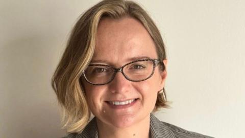 A woman with short blonde hair and glasses smiles at the camera. She is standing in front of a white wall. 