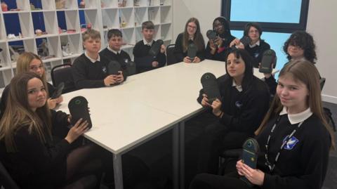 The photo shows a group of students looking at the camera. They are sitting around a table, and are wearing black jumpers, and are each holding a black pouch. There are white bookshelves behind them. 
