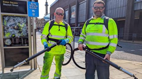 Two men wearing hi-vis clothing and gum removal backpacks. They are holding long poles attached to the backpacks and are standing on a "Bin Your Gum" sign on Newcastle's Blackett Street.