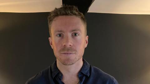 Frederick Fennessy from the shoulders up, he is wearing a blue button-up shirt and has short brown hair. The background is a blue wall.