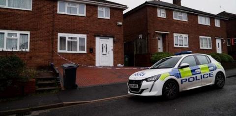 A police car is parked on the road outside a house, where police crime tape can be seen cordoning off the brick-paved driveway. The house has a white door and a white-framed front lower window.