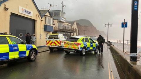 Police cars on a wet road with police tape up in Sidmouth. A HM coastguard is pictured too next to a lifeboat station.