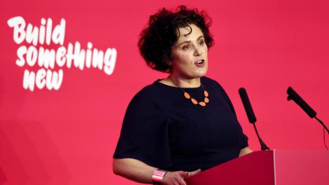 Claire Hanna, wearing a navy top, standing behind a red podium with two microphones on it. The wall behind her is read with "build something new" written in white. 