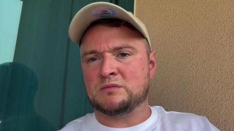 A man with a baseball cap in a white T shirt looks down the camera. He is sat on a hotel balcony.