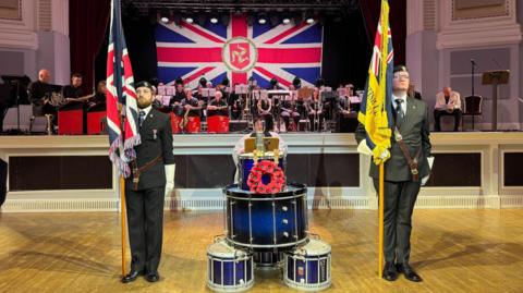 Two men standing in military uniforms and holding up flags n the Villa Marina Royal Hall. They are standing next to blue and white drums, which has a red wreath  been placed on one, and there is a large band sitting behind them.