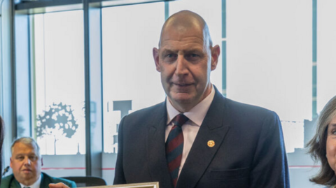 A man with a bald head in a dark blue suit jacket, pale shirt and blue and red tie