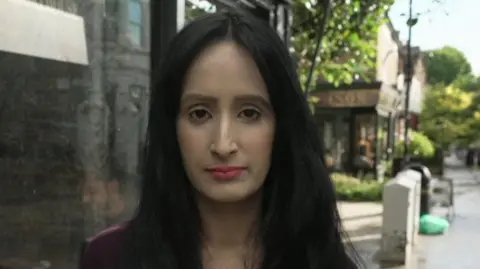 A woman with long dark hair stands on a high street outside a shop. The wet pavement and trees suggest recent rain, and a sign reading “The Axe and Ember” is visible in the background.