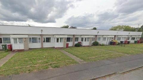 A row of 8 small, terraced bungalows is seen from the road via Google Maps. The doors and outside walls are white, each with a small square of mown grass in the front. They generally appear to be in poor condition. 