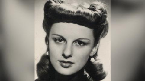 A black and white headshot image of Carol in her twenties looking down the lens of the camera. She is wearing drop white ball earrings and her hair is highly styled.