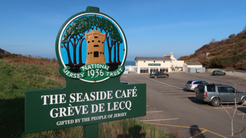 A large, bold, green 1936 Jersey national trust sign with a picture of a castle. The words 'The Seaside Cafe' can be seen with a moderately busy carpark in the background. The sky is blue with no clouds in the distance.