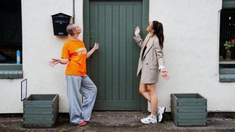 Two women standing in front of a doorway, posing with their arms out. The woman on the left is wearing an orange T-shirt and blue jeans and has a shaved head. The woman on the right is wearing a dress and a blazer and has long, dark hair tied back.