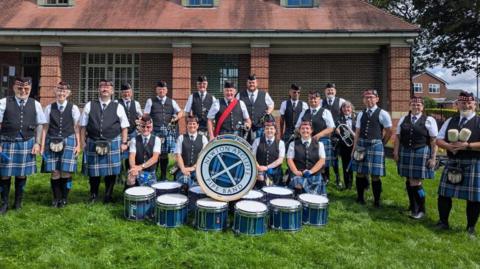 The members of the Newton Aycliffe Pipe Band are standing in a line with four members kneeling in front of them. A pile of blue drums, about the size of large tom-toms, are in front of them with a bass drum on top containing the logo and name of Newton Aycliffe Pipe Band in writing curving around the drumskin. The band members are wearing blue kilts and formal uniforms.
