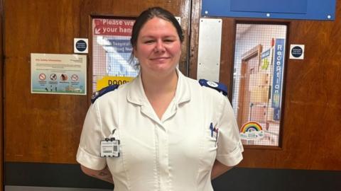 A woman in a white nursing uniform stands in front of doors to a hospital ward. Her hair is tied back and she smiles for the camera.