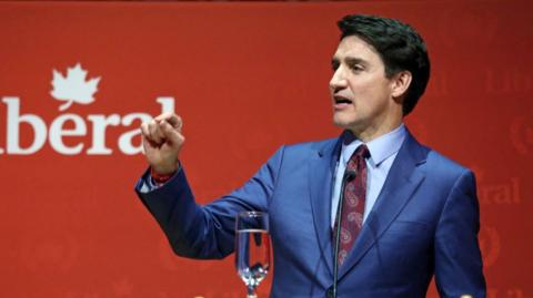 Justin Trudeau standing at a podium in front of a red back drop. He is wearing a blue suit and patterned tie and is pointing with one raised hand.