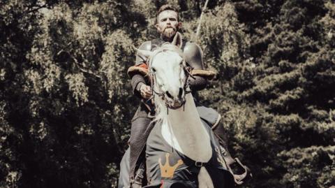 Henry Browne - a man dressed in a medieval knight uniform - has brown hair, a moustache and beard and is looking directly at the camera with a serious expression whilst he is sitting on a white horse. Trees can be seen behind him.