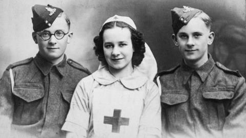 A grainy photograph showing three people. On either side are young men in army fatigues and cap. The man on the left also has a pair of thick-rimmed black glasses. In the middle is a woman with curly dark hair, wearing what looks to be a white nurses uniform, complete with cross on her chest.