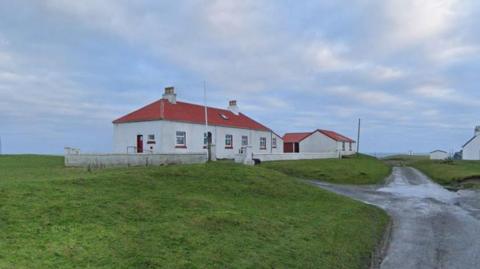 A white building with a red door and red roof. The building is surrounded by grass with a single track road running down the right hand side. The sky is dappled with clouds.