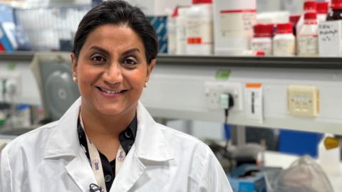 Dr Kris Subramaniam wears a white lab coat and stands in a lab with shelves filled with tubs and bottles behind her. She is wearing her dark hair tied back and has pearl earrings in. Underneath her lab coat she is wearing a dark coloured blouse and smiles at the camera.