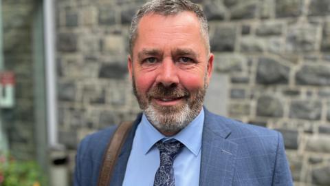 Deputy Marc Leadbeater - A man with black and grey hair looking at the camera. he also has a black and grey beard. He is wearing a blue checked suit, with a blue shirt and a blue patterned tie. Over his right shoulder he has a brown strap from a bag.