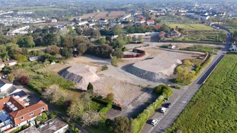Drone shot overlooking the Overdale Acute Hospital development site in St Helier, Jersey. Large mounds of building material are at the site. The area is surrounded by trees, homes and other buildings.