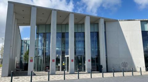 The picture shows the exterior of the court. It has a number of grey pillars resembling bars in front of a glass frontage. There is a solid grey wall with a royal crest.