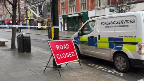 Police cordon tape is stretched across a deserted Oxford Street. In the foreground is a police forensic services van and a red sign saying "road closed".  