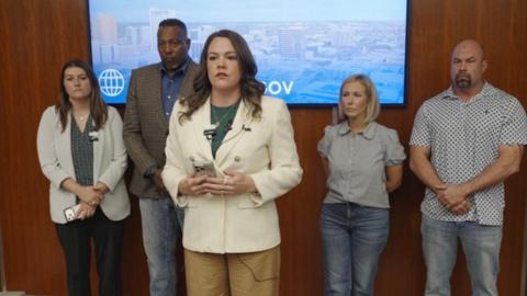 A woman in a white blazer stands in the foreground with a cell phone in her hands as she speaks. In the background of her are four other individuals leaning up against a wooden wall. 