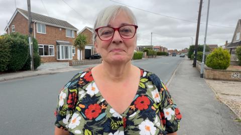 A woman wearing red glasses and a dark floral-patterned top stands in the middle of a residential street. Telegraph poles line both sides of the road, with detached houses, driveways and a cloudy grey sky in the background.