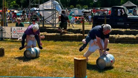 People hunch over grey metallic barrels and push them over grass. They are wearing sheep outfits around their legs.