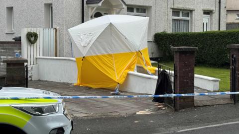The crime scene at Rocky Road Merthyr Tydfil. There's a forensic tent outside a white house with a white garden wall. A police car is on the left hand side and there's police tape in front of the house.