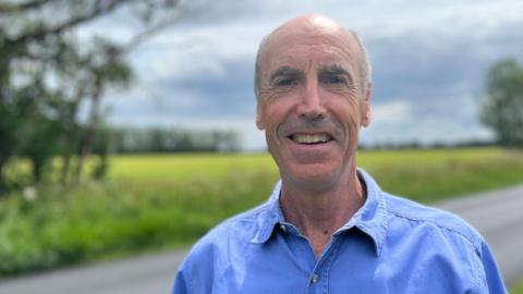 Head and shoulders image of Paul Boothman. Paul is wearing a blue shirt and is standing in front of a field and a road.