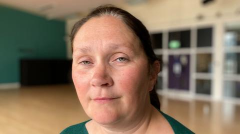 Det Ch Supt Linday Fisher - a woman with tied back dark hair wearing a green top stands in a room with a green wall to her left and a row of windows behind her. The background is blurred.